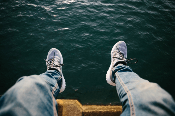 A man with his feet hanging off a pier wearing grey sneakers.