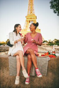 Two female friends sit on a ledge with the Eiffel tower in the background.