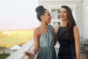 Two women in dresses share a laugh on the balcony of a building.