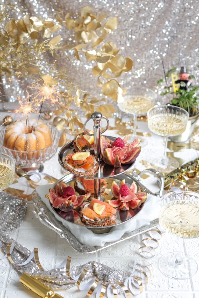 A two-tier platter of fruit is pictured sitting near several champagne glasses, with gold, sparkly decorations in the background.