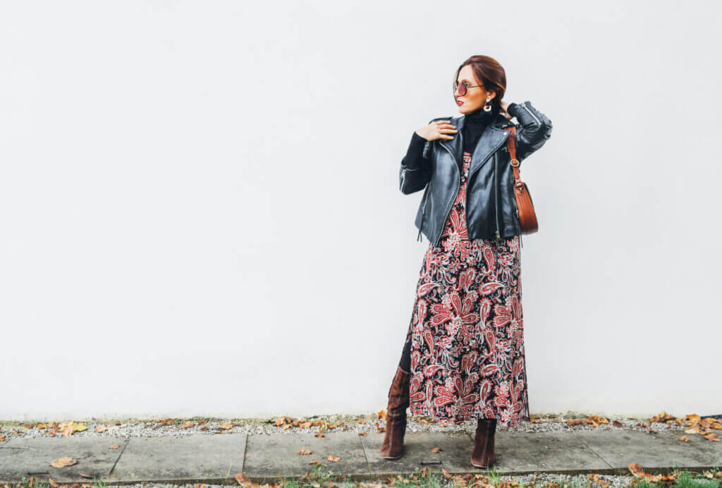 A woman is pictured wearing a patterned sundress, a moto jacket and suede boots.