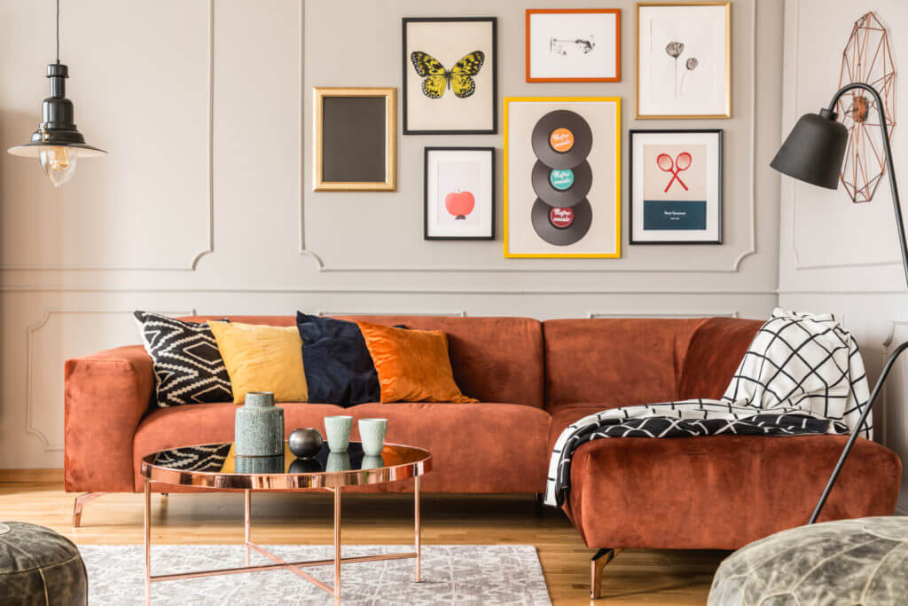 An orange couch is pictured in the corner of a room, with a series of framed pictures hanging on the wall above it.