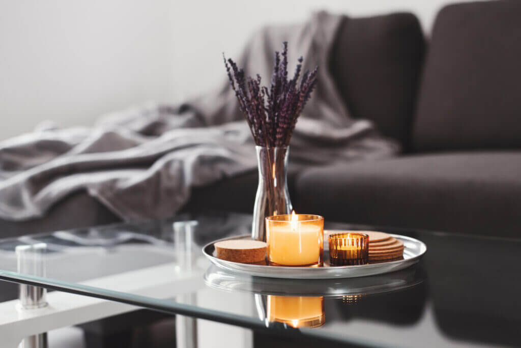 A tray holding two lit candles sits atop a glass table, next to a vase with flowers.