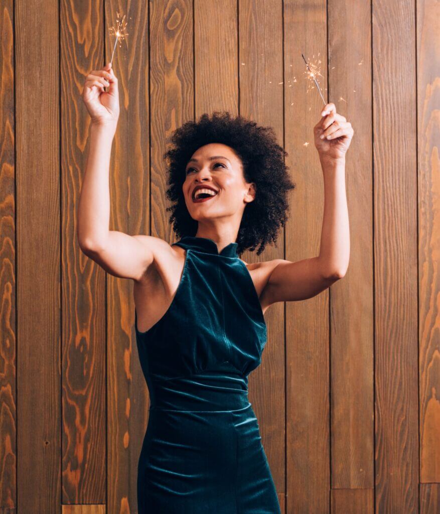 A woman in a green velvet dress smiles as she holds up two lit sparklers.