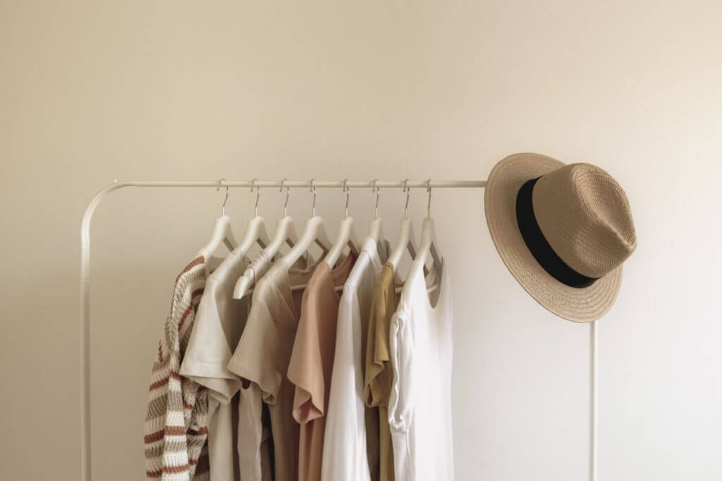A picture of a rack of clothing, with eight neutral-colored garments and a tan hat resting on the top frame.