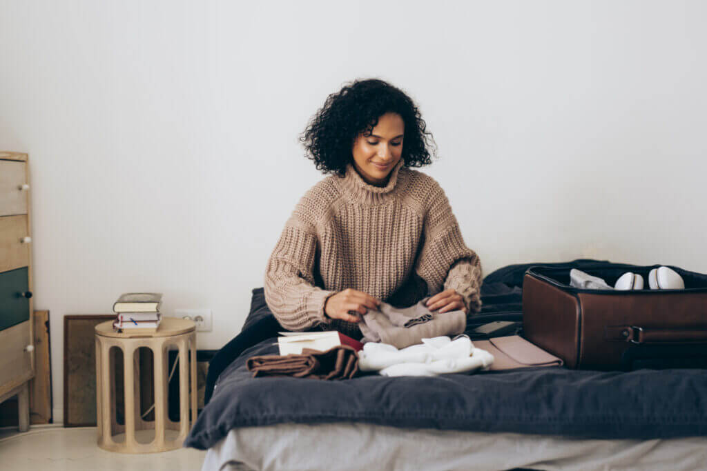 A woman sits atop her bed while wearing an oversized knit sweater.