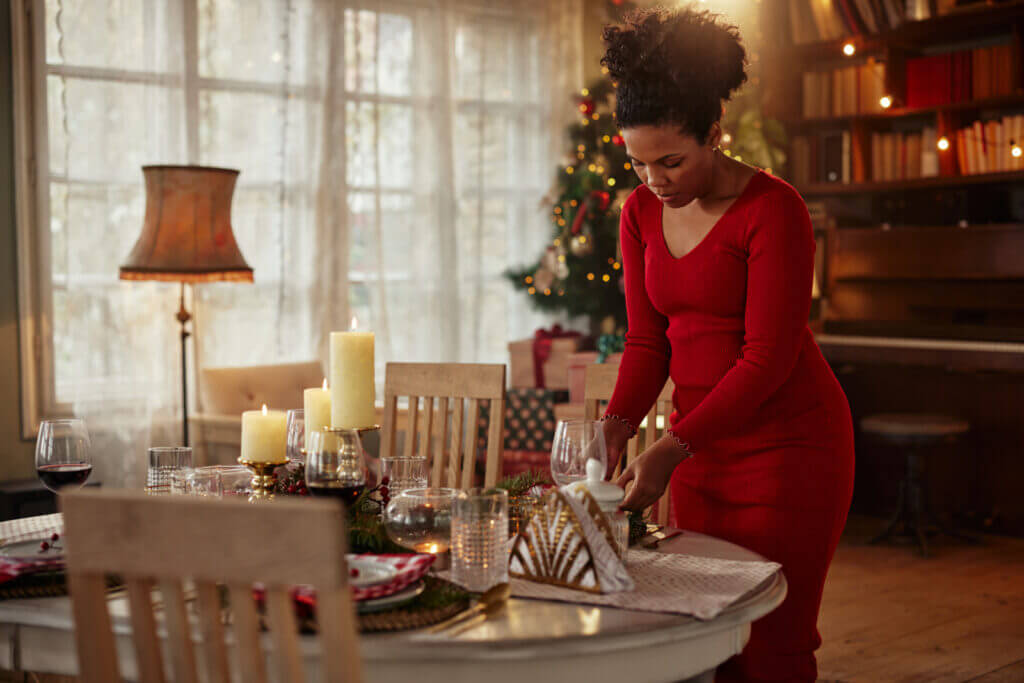 A woman in a red dress prepares the dining room table with place settings, with a Christmas tree lit up in the background.