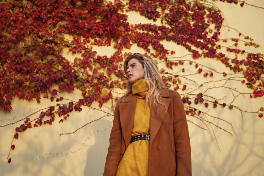 A stylish woman is pictured wearing a brown trench coat over a yellow sweater dress.