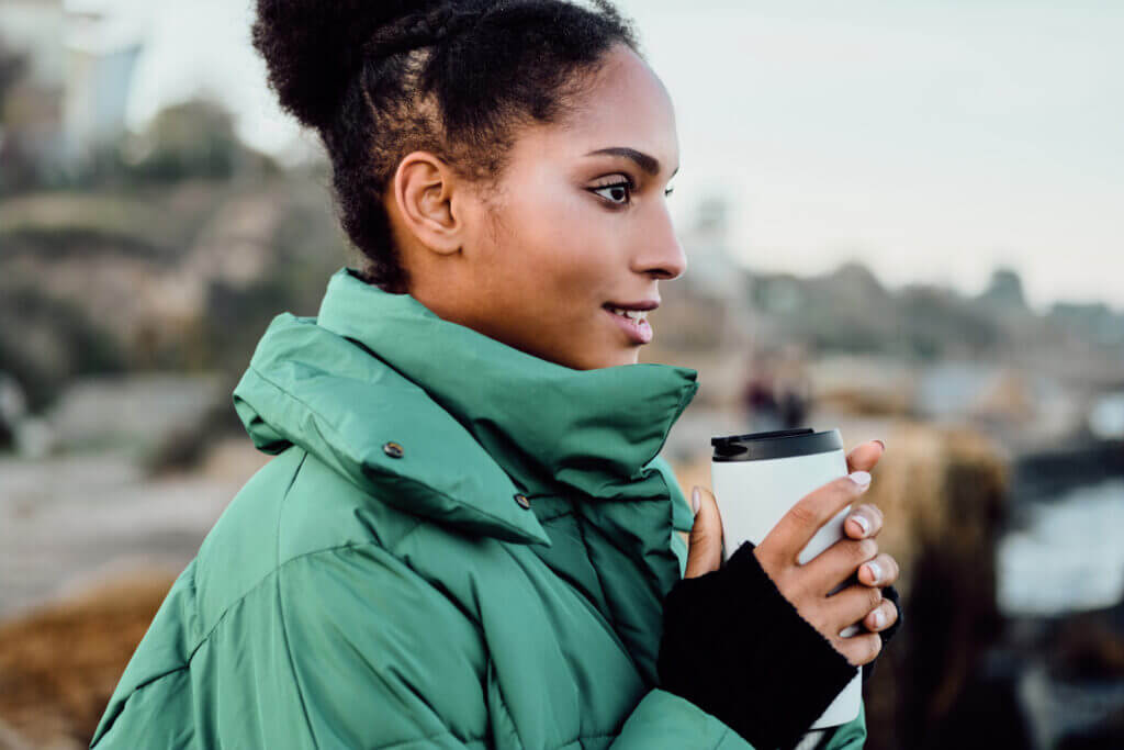 A woman in a green puffer jacket holds onto her coffee as she looks off into the distance.