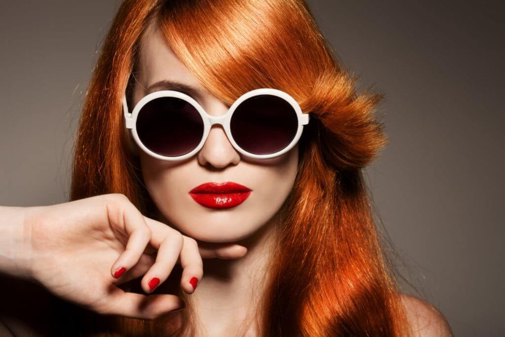 A close up picture of a woman with vibrant orange hair and large circular sunglasses with a white frame.