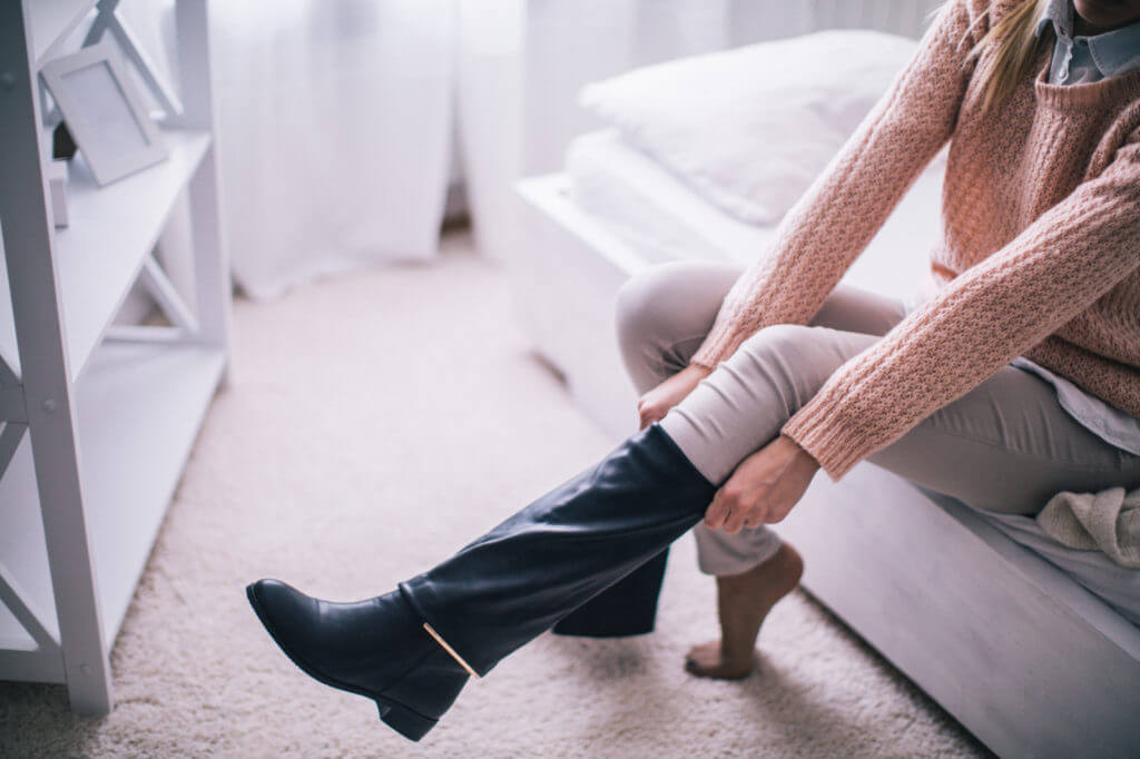 A woman wearing a knit sweater is pictured sitting on the side of a bed, putting on a knee-high boot.