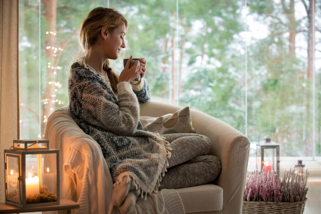 A picture of a woman cuddled up in a cozy chair, wearing a sweater while drinking a warm beverage.