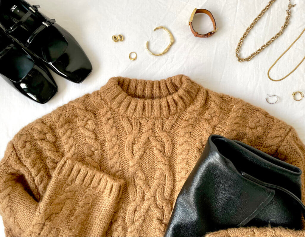 A gold cable knit sweater is laid out on a bed, with several accessories displayed nearby.